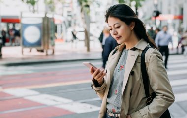 Cheerful pretty young woman in trendy wear walking on street with sunshine enjoying reading sms with good news on smart phone. happy asian lady holding cellphone standing on zebra crossing road.