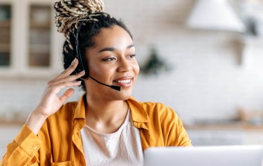 Joyful contented friendly young African American woman with dreadlocks and headphones, support operator, call center employee, working at laptop, advises clients online, looking away, smiling